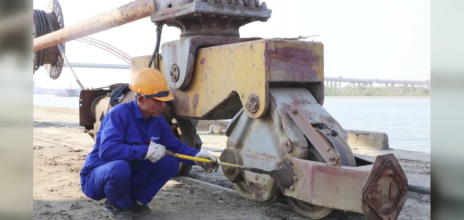 Công nhân đội mũ bảo hộ màu cam đang bảo dưỡng bánh xe cần cẩu cũ trong cảng Hoàng Diệu Hải Phòng, phía sau là sông Cấm và cầu Hoàng Văn Thụ