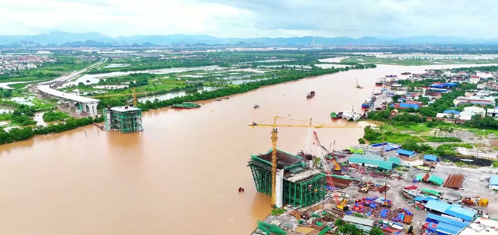 Toàn cảnh cầu Nguyễn Trãi đang thi công vượt sông Cấm Hải Phòng, hai trụ tháp mọc lên từ mặt nước, cầu dẫn bê tông chạy dọc bờ bên trái, tàu hàng qua lại giữa sông