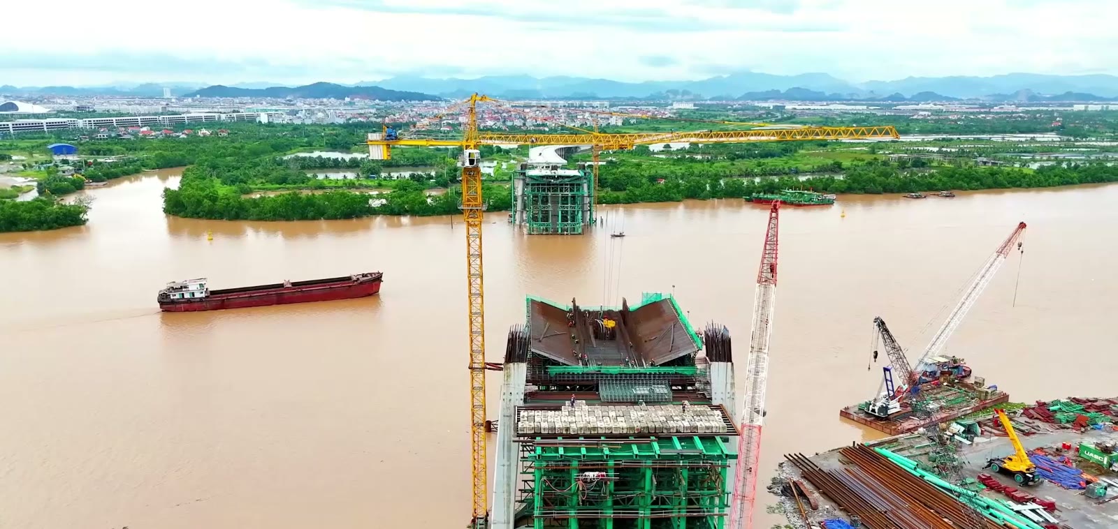 Trụ tháp cầu Nguyễn Trãi đang thi công giữa sông Cấm với giàn giáo màu xanh và cần cẩu tháp, một sà lan chở hàng chạy qua phía sau